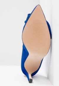 Escarpin en daim bleu à bout pointu avec une semelle beige et un talon aiguilles, photographié par le dessous.