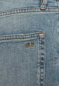 Ljust blå denimjeans med en ficka som har orange broderi som stavas "IVY." Tyget har ett synligt twillmönster och sömdetaljer.