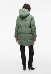 Piepildīta zaļa mētelis ar kapuci, ar horizontālu quilted rakstu. Valkāts virs melnām biksēm, redzams no aizmugures. Gluda tekstūra, vidēja garuma dizains.