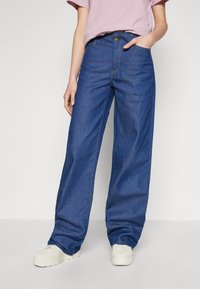 Dżinsy z wysokim stanem, szerokie nogawki w ciemnoniebieskim kolorze z widocznym przeszyciem, przednia kieszeń i zapięcie na guzik. Połączone z białymi trampkami.