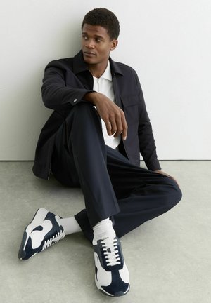 Jonge man zittend op de grond tegen een muur, gekleed in een zwart jack, wit overhemd, zwarte broek, witte sokken en zwart-witte sneakers.