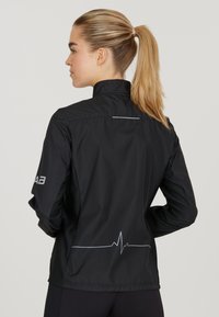 Schwarze Laufjacke mit hohem Kragen, dünnem weißen Herzschlag-Graphic auf dem Rücken und Branding am Ärmel. Leicht und mit glatter Textur.