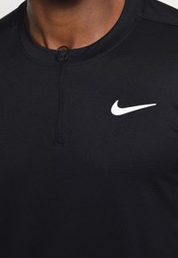 Musta urheilupaita, joka on sileää kangasta ja jossa on puolivetoketju kauluksessa sekä pieni valkoinen Nike-logo vasemmassa rinnassa.