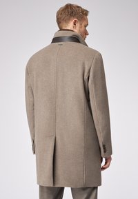 Cappotto di lana beige con colletto alto, tessuto testurizzato, spacco singolo sul retro e polsini abbottonati, che mostra un design minimalista.