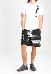 Weißes Kurzarm-Sportshirt mit grafischem Design, kombiniert mit schwarzen Shorts, die abstrakte weiße und graue Muster aufweisen. Schwarze Sportschuhe.