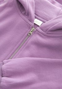 Gros plan sur un sweat à capuche zippé lavande montrant une fermeture éclair argentée et une étiquette blanche avec des détails de la marque et de fabrication.