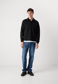 Fekete polo pulóver gallérral és elöl varrási részletekkel, világoskék farmernadrággal és fekete sneaker cipőkkel párosítva. Egyszerű háttér.