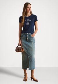 Μπλε βαμβακερό t-shirt με λευκό λογότυπο, φούστα από ανοιχτό μπλε τζιν με κουμπιά και σκίσιμο, καφέ ψηλοτάκουνα παπούτσια και μικρή καφέ τσάντα.