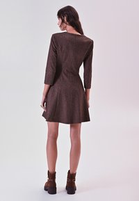 Bruine jurk tot de knie met driekwart mouwen, textuurpatroon, aansluitend lijfje, uitlopende rok en een ronde halslijn. Draagt het met een paarbruine enkellaarzen.