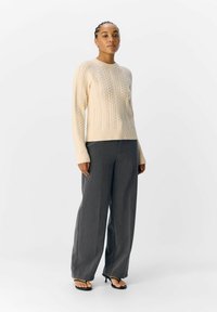 Krämfärgad stickad tröja med texturerad kabeldesign, tillsammans med grå vida byxor och svarta platta sandaler. Modellen står mot en enfärgad bakgrund.