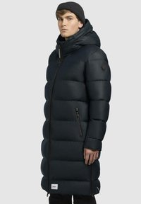 Sort pufferjakke med et langt, quiltet design. Har en høj krave, frontlynt, sidelommer og et logo patch på ærmet.