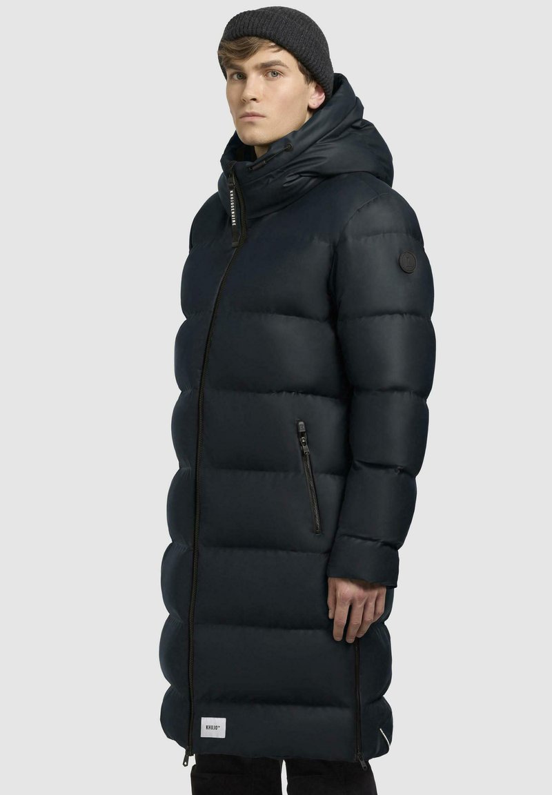 Sort pufferjakke med et langt, quiltet design. Har en høj krave, frontlynt, sidelommer og et logo patch på ærmet.