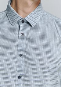 Chemise à manches courtes, col classique en bleu clair avec un motif géométrique. Présente deux boutons foncés, un tissu texturé et des coutures en contraste subtiles.