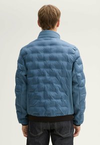 Puchowa kurtka w jasnoniebieskim kolorze, z fakturowanym pikowanym wzorem, stójką i czarnym ściągaczem na dole. Idealna na codzienne noszenie.