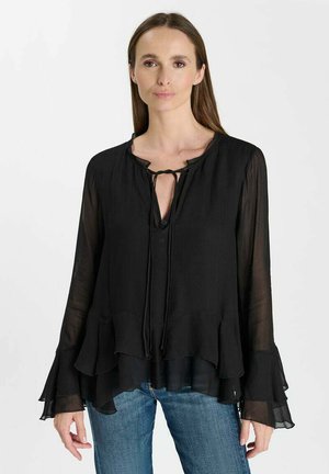 Vrouw met lang bruin haar die een zwarte doorschijnende blouse met geplooide manchetten en zoom draagt, gecombineerd met een blauwe spijkerbroek, staand tegen een witte achtergrond.