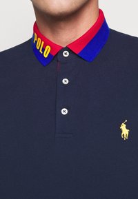 Sjöblå pikétröja med en röd och blå ribbad krage. Har gul "POLO" text, tre knappar och en liten broderad gul logotyp.