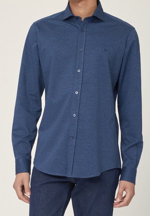 Hombre con una camisa azul ajustada de manga larga con un sutil patrón de puntos y vaqueros azul oscuro, de pie frente a un fondo liso.