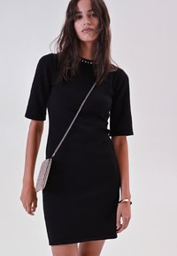 Abito nero a maniche corte realizzato in tessuto morbido, con un colletto rotondo arricchito da borchie argentate. Indossato con una piccola borsa a tracolla decorata.