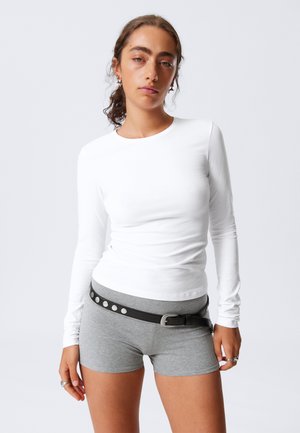 Camisa branca de manga longa combinada com calções cinzentos. Os calções têm um cinto preto com tachas prateadas. Tecido de textura suave; design justo.