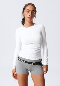 Chemise blanche à manches longues associée à un short gris. Le short est doté d'une ceinture noire avec des clous en argent. Tissu au toucher lisse ; design ajusté.
