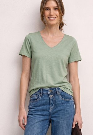 Młoda kobieta uśmiecha się, ubrana w jasnooliwkowy T-shirt z dekoltem w serek i niebieskie dżinsy, stoi na tle jednolitej jasnej ściany.