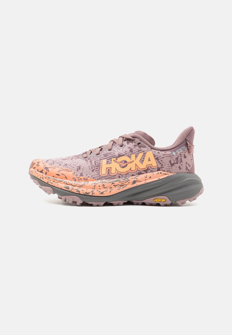 Terrengløpesko med en teksturert lavendeloverdel, oransje detaljer og robust grå yttersåle. Har en dristig logo og Vibram-merke.