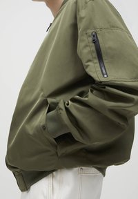 Oliwkowo-zielona kurtka bomberka z kieszenią na zamek, ściągaczami przy mankietach oraz luźnym krojem. Gładka, matowa tkanina z detalami przeszyć.