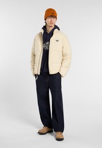 Giacca a vento color crema, con chiusura a zip e tasche laterali, indossata sopra un felpa blu navy, abbinata a jeans blu scuro e stivali color cammello.