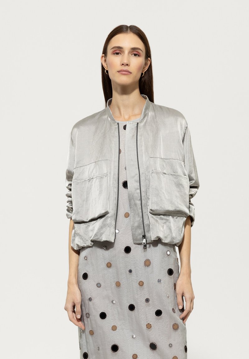 Silver bomber jacket in lightweight material, featuring a zip front, two large front pockets, and relaxed fit, paired with a dotted dress.