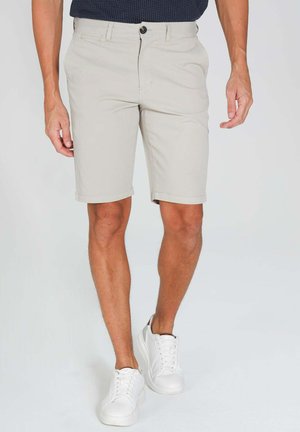 Homme portant un short beige clair et des baskets blanches, debout les mains détendues le long du corps devant un fond uni.
