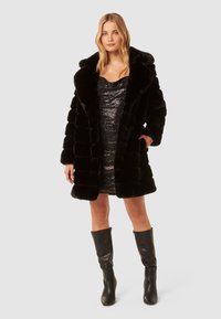 Donna che indossa un cappotto di pelliccia sintetica nero fino al ginocchio sopra un miniabito nero decorato con paillettes e stivali neri alti fino al ginocchio, in piedi su uno sfondo semplice.