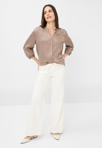 Blouse en soie marron clair avec un décolleté en V et des poignets élastiques, associée à un pantalon large blanc et des ballerines beiges pointues, sur un fond uni.