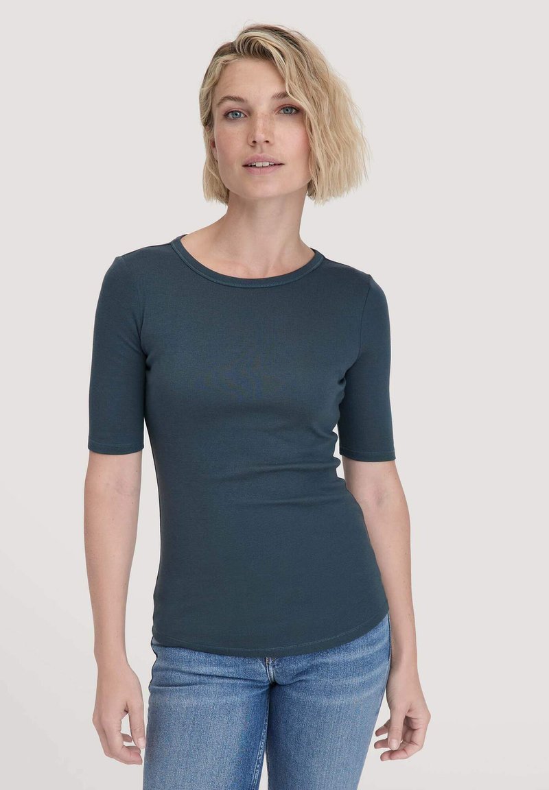 Strak, kortmouw teal shirt met een ronde halslijn en glad, rekbaar materiaal. Gecombineerd met lichtblauwe jeans.