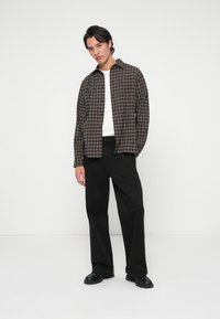 Camicia di flanella a quadri in tonalità scure, abbinata a pantaloni larghi neri. Il modello sta in piedi, mostrando un taglio rilassato e un design semplice.