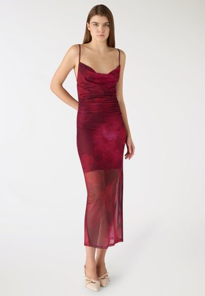 Giovane donna in piedi che indossa un vestito lungo senza maniche trasparente di colore rosso scuro con tacchi beige appuntiti su sfondo bianco.