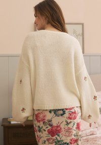Maglione lavorato a maglia color crema con maniche ampie, decorato con ricami floreali sui polsini. Indossato sopra una gonna con motivo floreale.