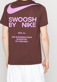 Brązowa bawełniana koszulka z dużym różowym napisem "SWOOSH BY NIKE" oraz logo na plecach. Prosty okrągły dekolt i krótkie rękawy.
