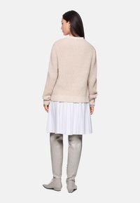 Beige gestrickter Pullover mit langen Ärmeln, gerippten Bündchen, über einem weißen Faltenrock getragen, kombiniert mit grauen kniehohen Stiefeln.