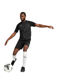 Zwarte sportieve Puma-shirt met een gestructureerd patroon, zwarte shorts en witte sokken; speler dribbelt een zwart-wit voetbal.