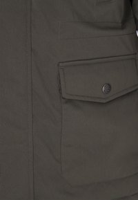 Giacca in tessuto texturizzato verde scuro con una grande tasca con bottone a pressione e dettagli di cucitura sottili. Design casual con elementi funzionali.