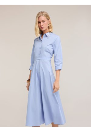 Frau trägt ein hellblaues Midi-Kleid mit Dreiviertelärmeln, tailliertem Gürtel und verziertem Kragen, steht vor einem beigen Hintergrund.