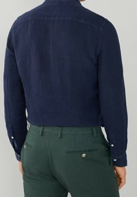 Camicia di lino blu navy con maniche lunghe e polsini con bottone, abbinata a pantaloni verdi con tasche posteriori e dettagli con bottoni.
