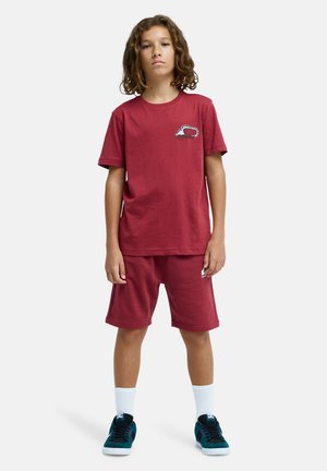 T-shirt en coton bordeaux à manches courtes avec un détail de logo, associé à un short assorti. Porté avec des chaussettes blanches et des baskets noires et bleu-vert.