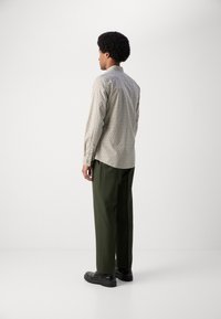 Beige színű, geometrikus mintázatú ing, sötétzöld nadrággal párosítva. A modell háttal áll a kamerának, fekete cipőt visel.