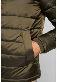 Olivově zelená péřová bunda s prošitou strukturou, vybavená boční kapsou a žebrovaným límcem. Materiál působí hladce a mírně leskle.