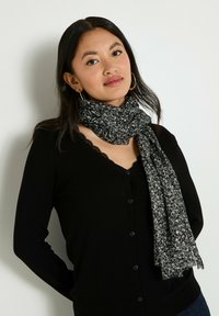 Écharpe florale noire en tissu doux, présentant un motif texturé avec des accents blancs et jaunes, tombant autour du cou sur un haut noir.