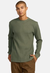 Langärmliges, olivgrünes Waffelstrickshirt mit Rundhalsausschnitt und Rippstruktur. Verfügt über ein dezentes Logoetikett nahe dem unteren Saum.