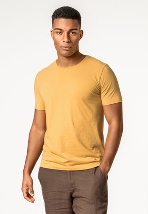 Junger Mann mit kurzen lockigen Haaren, der ein senfgelbes T-Shirt und braune Hosen trägt, steht mit einer Hand in der Tasche vor weißem Hintergrund.