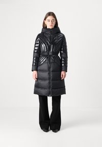 Čierna puff coat s lesklým povrchom, opasok v páse, stojatý golier a horizontálne prešívanie. Nosené cez čierne rozšírené nohavice.