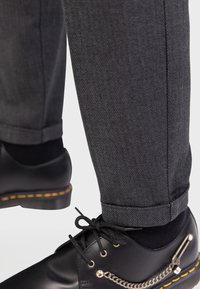 Chaussures en cuir noir avec détail en chaîne dorée assorties à un pantalon à motifs en chevrons gris avec des ourlets et des chaussettes noires sur un fond blanc.